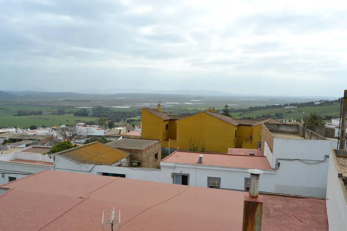Whitewashed rooftops of a hill town in the foreground and a wide green plain stretching to a hazy horizon under a pale sky